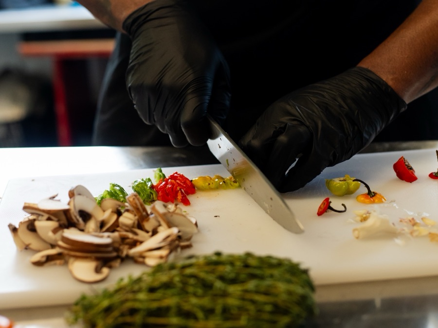 A chef in black gloves slicing scotch bonnet peppers on a board beside mushrooms and fresh thyme