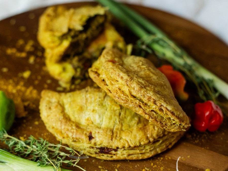 Two golden patties on a wooden board with green onion, thyme and a scotch bonnet pepper